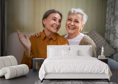 Two elderly sisters having fun indoors. Cheerful senior woman enjoying nice time together with her best female friend, sitting on couch at home, laughing, telling jokes. People, aging and friendship Wall mural