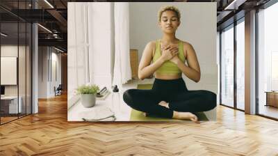 Young African American lady practicing yoga at home in black leggings sitting on floor in lotus posture hands crossed on chest slight delight smile on face, feeling her body. Emotional health Wall mural