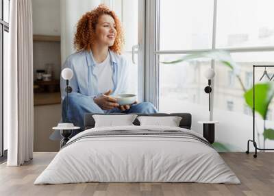 Young woman with red curly hair in jeans and shirt sitting on window sill looking through big window on city holding bowl of breakfast in hands, smiling noticing something funny outside Wall mural