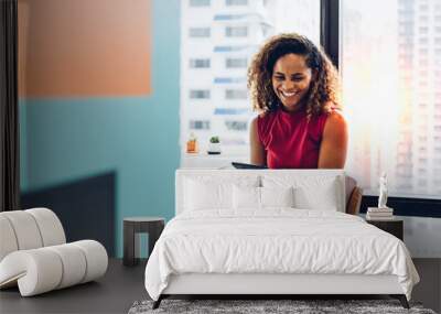 Portrait of cheerful black skin african woman smiling and using tablet for video conversation while relaxing on desk in modern office. Concept of young business people working at home. Wall mural