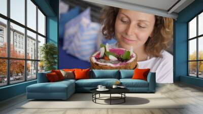 Curly girl looks closely with excitement at tropical smoothie bowl decorated with berries and toasted coconut chips served in coconut shell Wall mural