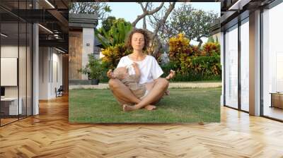 Young Caucasian woman breastfeeds her baby daughter while sitting in lotus position in a garden  Wall mural