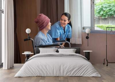 A smiling young Asian female nurse having fun talking with an Asian male cancer patient in a head cover sitting on a wheelchair, while taking him to relax with green nature outside the open window. Wall mural