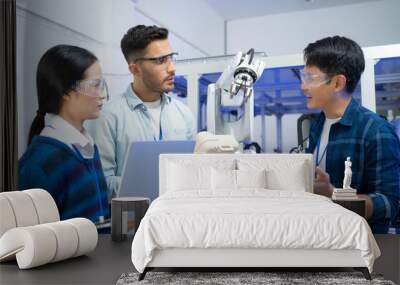 Caucasian male engineer standing hold a robot controller next to Asian female apprentice holding a laptop, demonstrating the work of robotic machine to young Asian male CEO who smile touching a robot. Wall mural