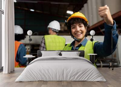 Selective focus, front view of a happy pretty short-haired Asian female factory worker in a vest and helmet, showing thumbs up while smiling at camera with blurred workers working in the background. Wall mural