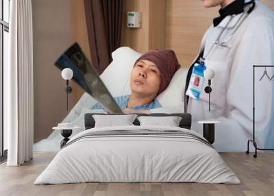 Selective focus of a depressed Asian male cancer patient in a head cover laying on bed looking at his MRI scan film in a cropped blurred female doctor's hand while diagnosing the film beside his bed. Wall mural