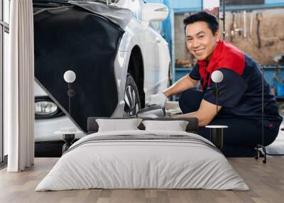 Selective focus of a mid-adult Asian male mechanic in uniform and gloves, squatting using a pneumatic wrench changing lug nuts of a car front wheel while turning to smile at the camera in the garage. Wall mural