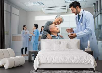 Selective focus of a smiling Caucasian male doctor standing holding hand while talk to a happy senior Asian male patient sitting on wheelchair pushed by his wife at a hospital. Copy space on left side Wall mural