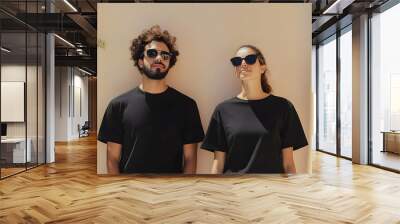 A man and woman, both wearing black t-shirts and sunglasses, stand side by side against a beige wall. Wall mural