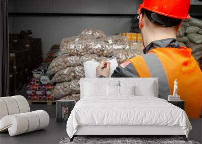 warehouse worker in a protective helmet and overalls checks the goods walnuts and dried fruits in the warehouse. warehouse logistics cargo courier, shipment transportation, worker holding clipboard Wall mural