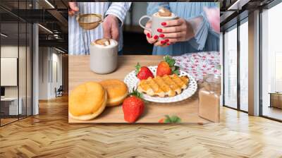 A mans and a woman's hands in light blue clothes with red nails. A man ads cinnamon in a gray cup with caffe latte. The crispy waffle, red strawberries on a white plate and donuts. Wall mural