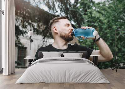 A handsome bearded guy 30 years old drinks water from a special plastic bottle on the street. Blue sports water bottle Wall mural