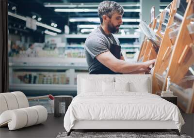 Busy male salesman is putting bread on shelves in bakery department in food store, bearded guy is wearing apron. Selling products, profession and trade concept. Wall mural