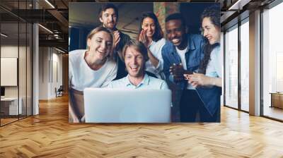 Excited young man is looking at laptop screen, rejoicing and expressing happiness, his happy colleagues are congratulating him on success, clapping hands Wall mural