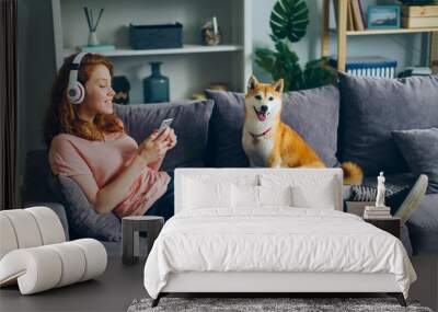 Joyful female student in headphones is listening to music and using smartphone sitting on couch with adoranle well-bred dog. People and modern lifestyle concept. Wall mural