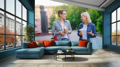 Joyful friends mature women are walking in street holding to go coffee and bright paper shopping bags talking enjoying conversation. Lifestyle and friendship concept. Wall mural