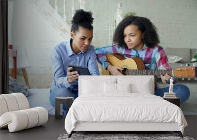 Mixed race young woman with tablet computer sitting on bed teaching her teenage sister to play acoustic guitar at home Wall mural