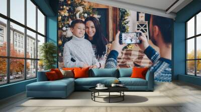 Mother and son are posing for smartphone camera near Christmas tree while father is taking photo, family is celebrating winter holiday. People and events concept. Wall mural