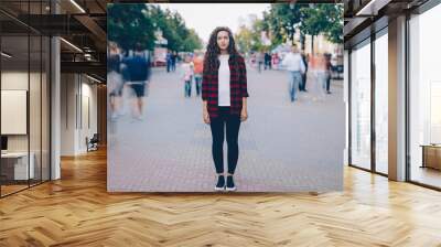 Portrait of beautiful young girl with long curly hair standing in street looking at camera when many men and women are walking around in hurry on summer day. Wall mural