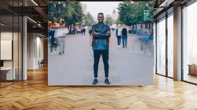 Portrait of happy African American guy wearing stylish jeans and leather jacket standing alone in street downtown, smiling and looking at camera with crowd moving by. Wall mural