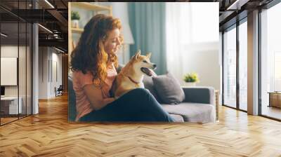 Pretty redhead woman is hugging cute doggy sitting on couch in apartment smiling enjoying beautiful day with beloved animal. People and pets concept. Wall mural