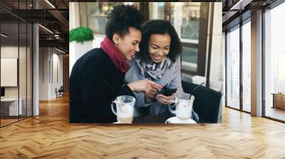 Two attractive mixed race female friends sharing together using smartphone in street cafe outdoors Wall mural