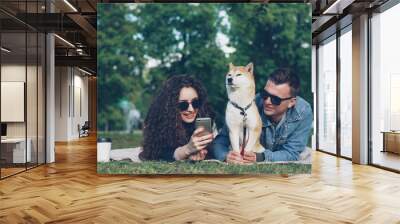 Young woman is showing funny pictures on smartphone to her cheerful boyfriend while relaxing in park with pet dog, people are lying on plaid on grass and watching screen. Wall mural