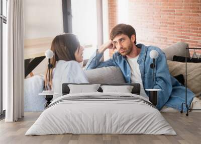 A young 25-year-old couple sits on their cozy sofa in a living room, engaged in a quiet conversation and sharing carefree moments together. Leisure and relaxation concept. Discussion man and woman Wall mural