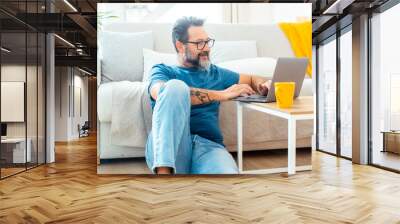 Adult man in casual clothing and eyeglasses using laptop and smiling while sitting on the floor against the sofa at home. Indoor technology computer leisure activity people lifestyle. Working online Wall mural