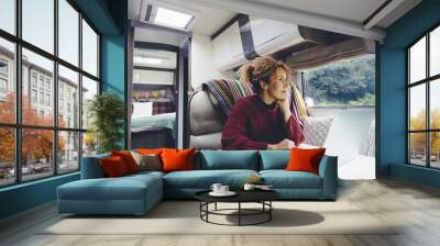 Adult woman use laptop computer inside a camper van recreational vehicle sitting at the table with bedroom in background and nature park outside the windos. Concept of travel and remote worker people Wall mural