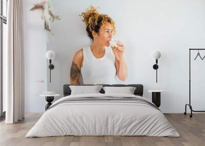 Attractive curly blonde middle age cute woman drinking water and lemon at home with white wall in background - healthy lifestyle for cacuasian people - modern tatoo girl Wall mural