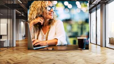 Attractive woman smile and look around while work on laptop sitting at the bar. Modern female people working online. Digital traveler nomad lifestyle. Happy female with glasses and notebook computer Wall mural