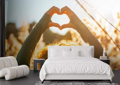 Back view of woman doing heart gesture with hands in front of an amazing golden sunset in the middle of high grass yellow field. Autumn season. Love nature. Concept of wellbeing. Serene people outside Wall mural