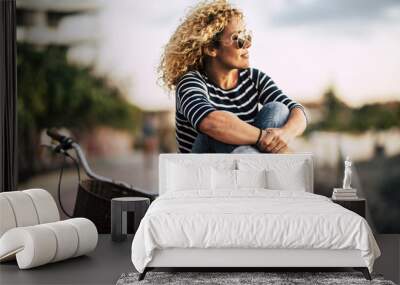 Beautiful adult caucasian young woman with blonde curly hair enjoy the outdoor leisure activity relaxing and sitting on a wall with bike in background - people portrait with sky in background Wall mural