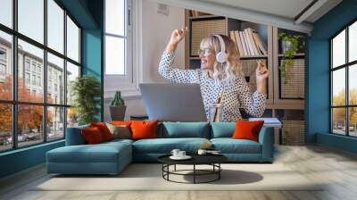 Businesswoman listening music on headphones while working on laptop at home office. Caucasian young woman enjoying music on headphones while looking out through window. Joyful woman with laptop  Wall mural