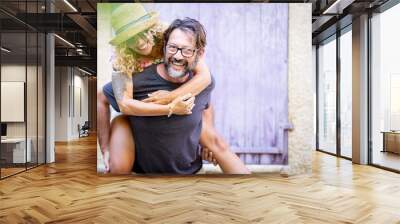 Carefree couple spending leisure time together outdoors, man giving piggyback ride to cheerful tattooed woman in sunglasses and straw hat. Woman enjoying piggyback ride on the back of her husband Wall mural