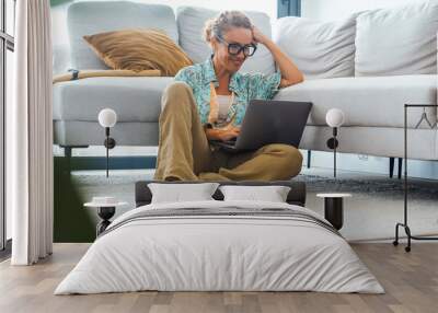 Carefree modern adult woman using laptop at home sitting on the floor against a grey sofa. Female people surfing the net with a computer alone in apartment having fun. Online activity leisure indoor Wall mural