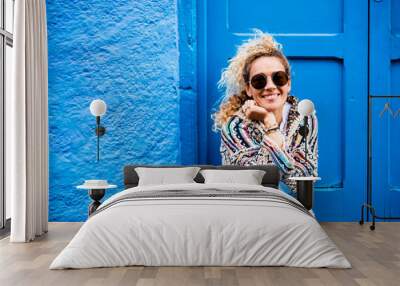 Cheerful adult beautiful woman portrait in the blue color - happy people sit down outside home and enjoy time - pretty young female with blue door and wall in background Wall mural