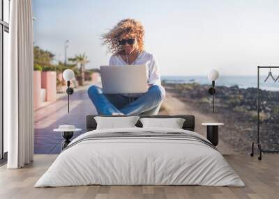 Cheerful beautiful blonde curly people young adult woman sitting outdoor with sea and sky in background working with tablet computer internet connected - freedom from office concept Wall mural