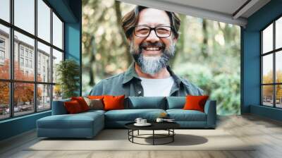 Cheerful portrait of bearded mature man looking on camera with green nature background. People enjoy outdoor leisure activity alone. Tourist at the park. Happy adult male smile. Front view person Wall mural