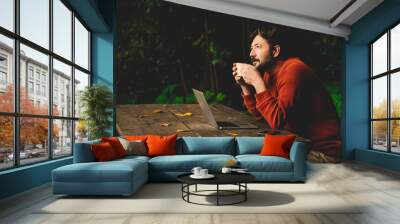 Concept of remote worker and smart working digital modern people lifestyle with adult hipster bearded man work outdoors on a table in the woods and drinking coffee thinking about job - online laptop Wall mural