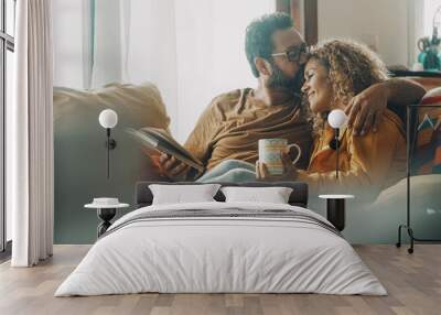 Couple of young adult man and woman enjoy time together sitting on the sofa in indoor leisure activity.. Real life people at home. Male and female relax on the couch reading a book and drinking a tea Wall mural