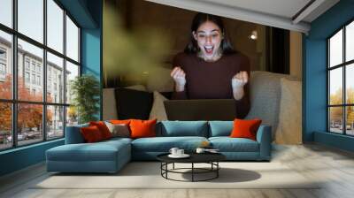 Excited young woman with a surprised expression looks at her laptop screen while sitting alone on a sofa at night. She is thrilled and happy, browsing the internet at home, celebrating  achievement Wall mural