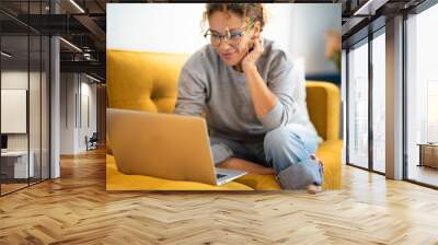 Female sitting on a yellow couch using laptop and internet connection and smile. Happy woman in indoor technology leisure activity. Social media life account concept. Adult Lady writing on computer Wall mural