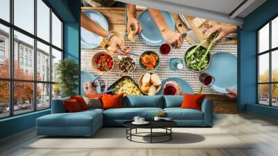 Group of friends eating together with glass of wine on dining table. Hands taking food from bowl on plate. Group of people enjoying variety of food and drinks at a getogether party Wall mural