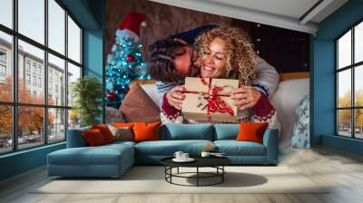 Happiness in christmas holiday eve season. Man doing a surprise to happy woman giving her a xmas gift in a red decorated box. Christmas tree in background. Couple celebrating together with love joy Wall mural