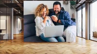 Happy adult aged couple caucasian people at home with personal laptop computer and internet connection - together in indoor leisure activity with technology sitting on the sofa and smiling Wall mural