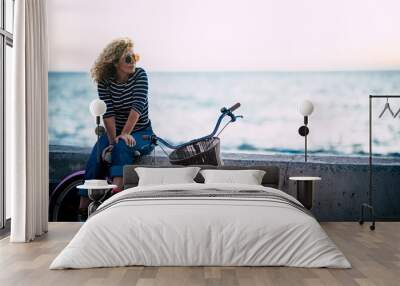 Happy beautiful curly healthy caucasian adult middle age woman sit down on a vintage coloured bike and enjoy the ocean in outdoor leisure activity - trendy people with hapy life Wall mural