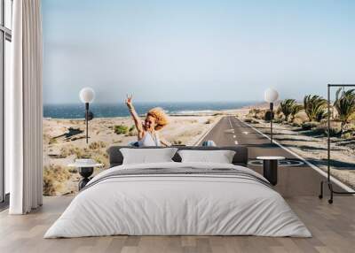 Happy cheerful couple of young caucasian crazy woman with curly hair enjoy the travel and vacation staying outside the roof of a convertible car with long road and ocean in background - joyful Wall mural