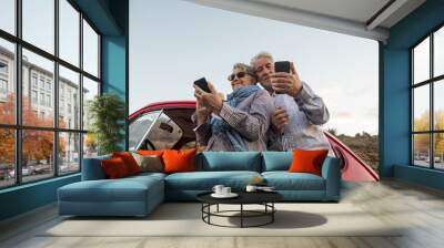 Happy elderly senior couple use mobile phones outdoor near a red beautiful retro car. leisure activity and travel together forever concept. modern use of technology from old adult that enjoyed life Wall mural